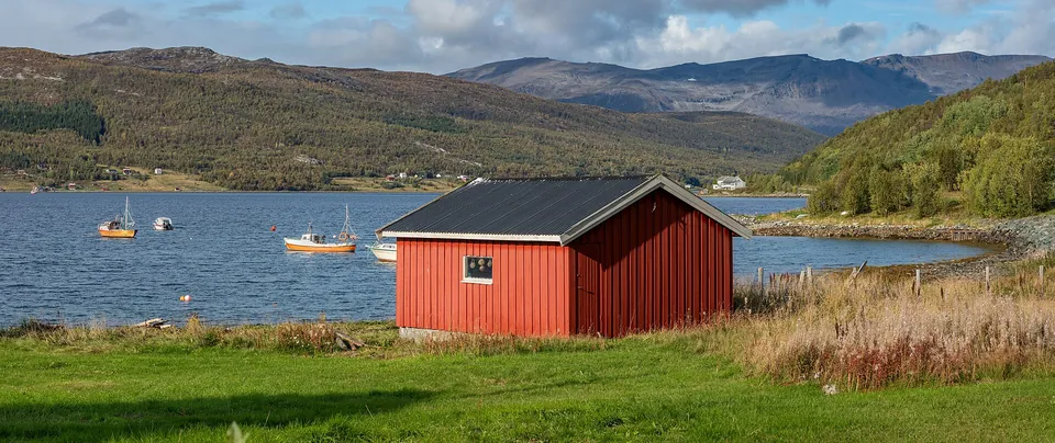 Rødt sjøhus og båter langs Burfjorden i Burfjord, med fjell og høstfarger i bakgrunnen.